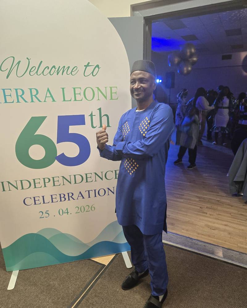 Abdullah Kamara, President of SALDAC, in a blue embroidered outfit and cap beside the Sierra Leone 65th Independence Celebration banner